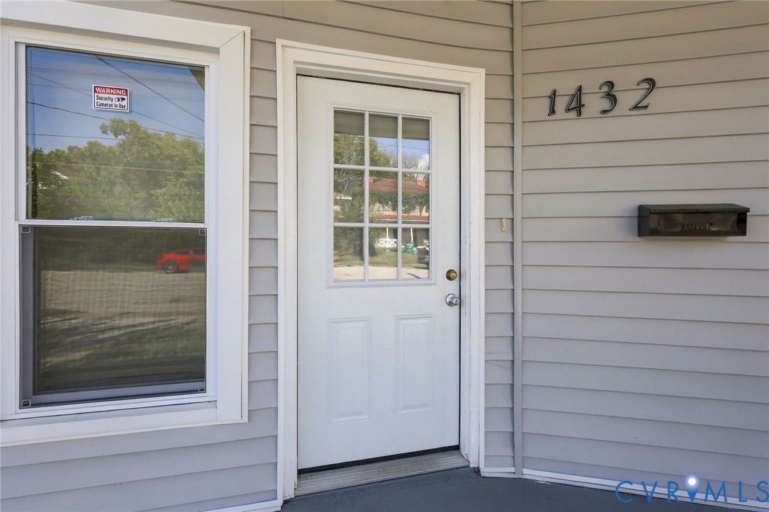 White paneled door with divided lite window, gray horizontal siding, and dark-finish house numbers