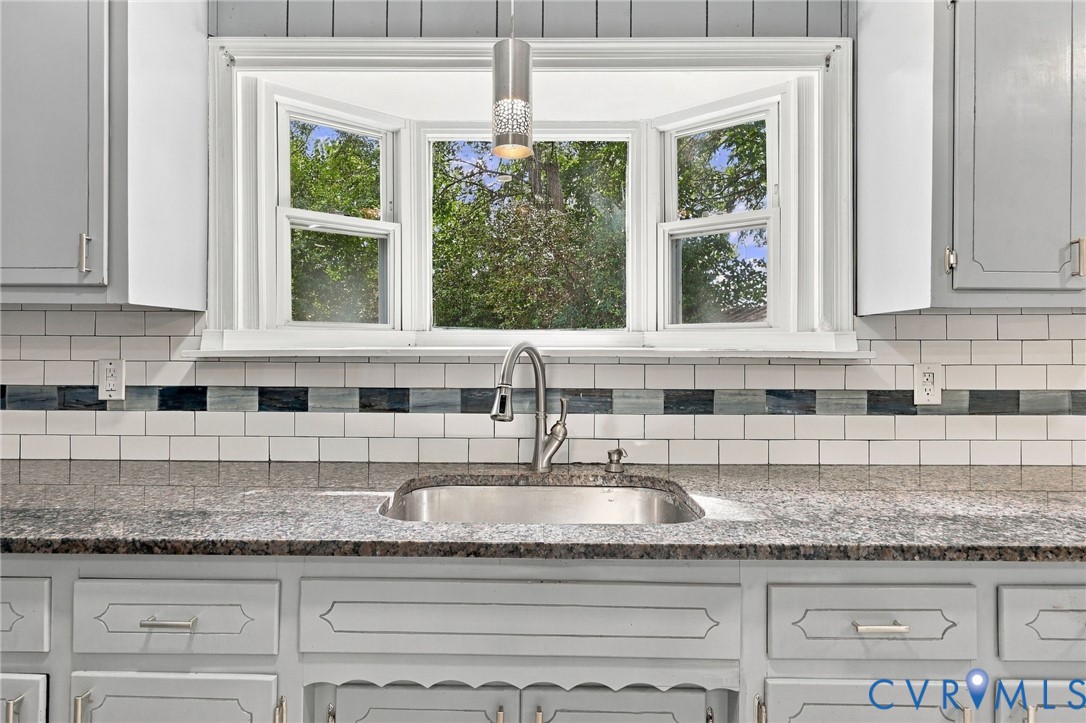 Bay window with white trim, undermount stainless steel sink, gooseneck faucet, light gray countertops, and subway tile backsplash with decorative accent