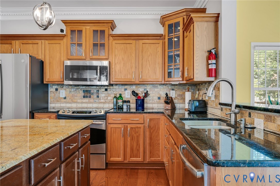 Chef's kitchen featuring warm wood cabinetry with glass-front upper cabinets, stainless steel appliances including a gas cooktop, dual granite countertops with waterfall island seating, and a professional-style faucet with pull-down sprayer