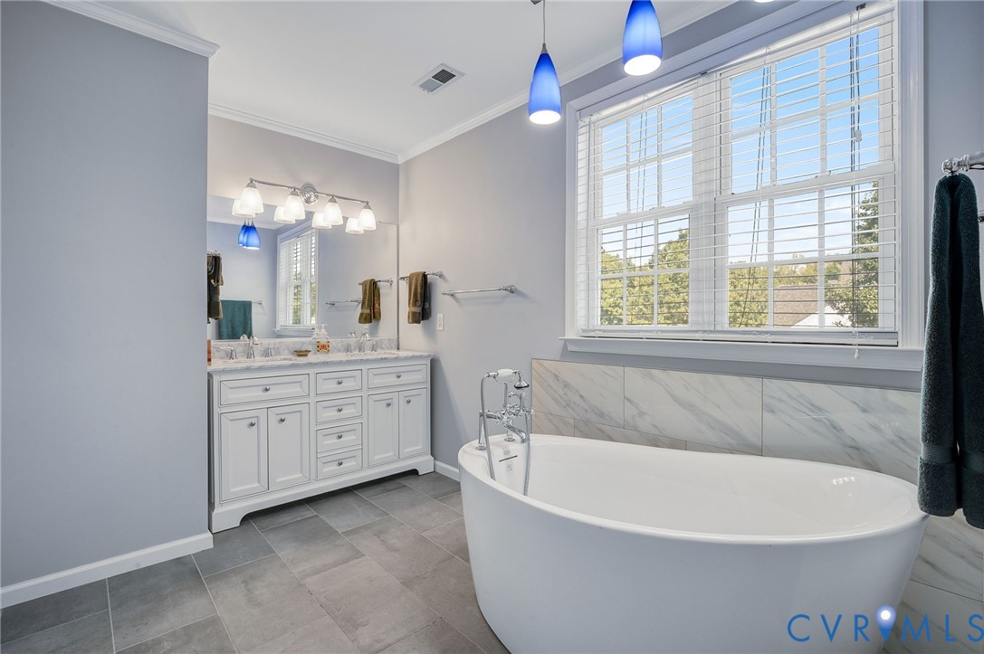 Freestanding soaking tub with a wall-mounted faucet, double vanity with stone countertop, large window with blinds, and tile flooring