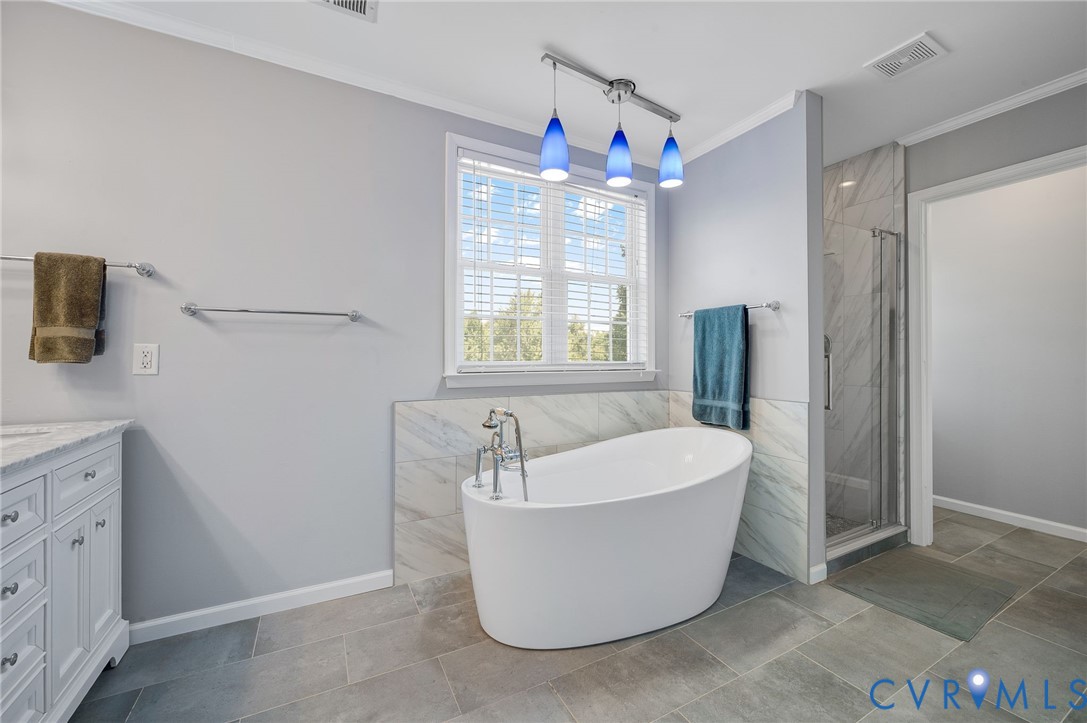 Freestanding soaking tub with floor-mounted fixtures, glass-enclosed shower with tiled surround, single vanity with light-colored countertop, and large format floor tiling