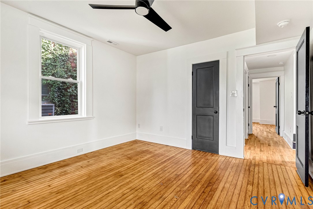 Hardwood flooring, white walls, black ceiling fan, window with exterior foliage, and a dark-paneled door