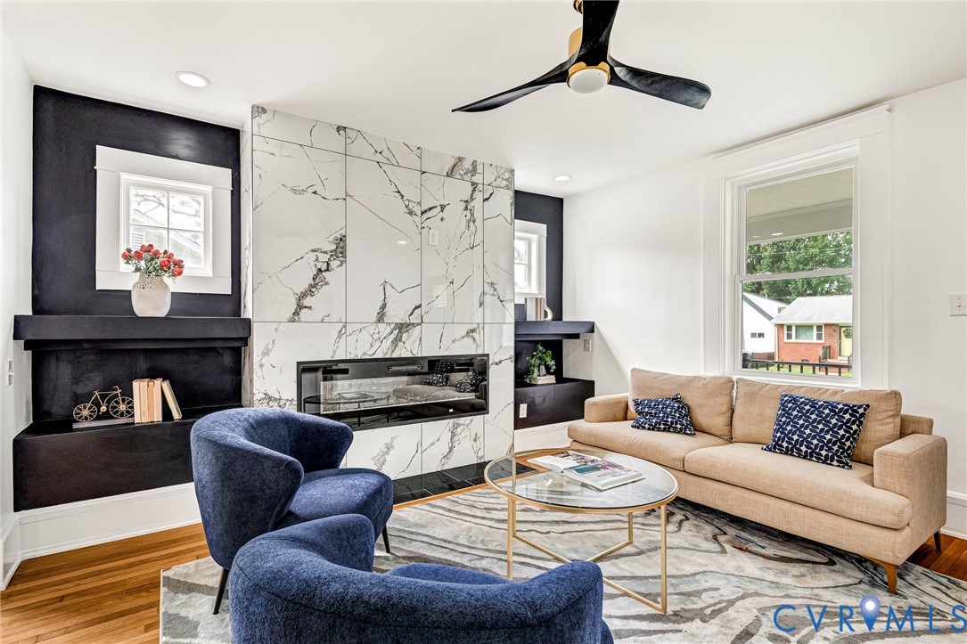 Integrated electric fireplace, large format marble-look tile surround, built-in shelving, wood-finish flooring, and a modern ceiling fan
