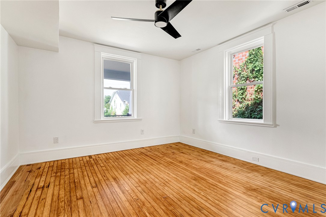 Hardwood flooring with a natural finish, white painted walls, two windows with white trim, and a black ceiling fan