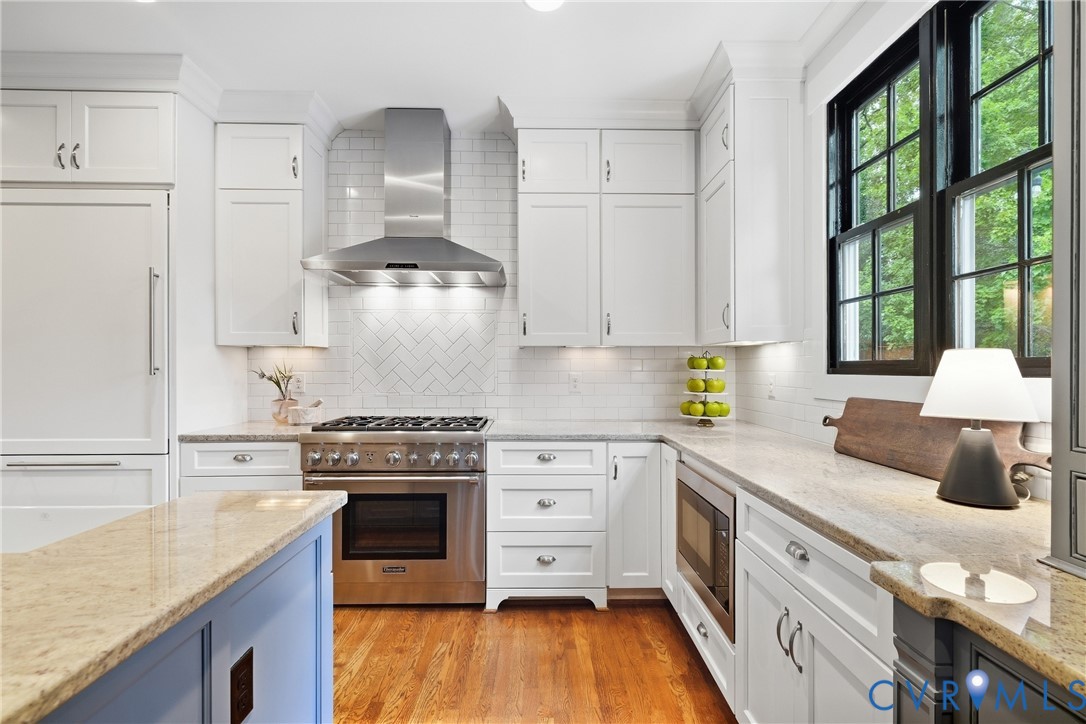 White shaker cabinetry with undermount lighting, stainless steel appliances, herringbone tile backsplash, light-toned granite countertops, and wood-finish flooring White shaker cabinetry with undermount lighting, stainless steel appliances, herringbone tile backsplash, light-toned granite countertops, and wood-finish flooring