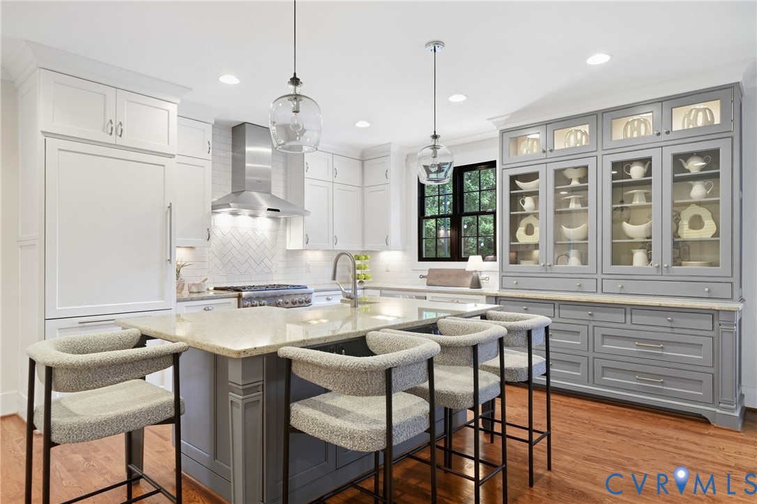Kitchen featuring a central island with countertop seating, white shaker cabinetry, a stainless steel range hood, a herringbone tile backsplash, and a built-in display cabinet with glass doors Kitchen featuring a central island with countertop seating, white shaker cabinetry, a stainless steel range hood, a herringbone tile backsplash, and a built-in display cabinet with glass doors