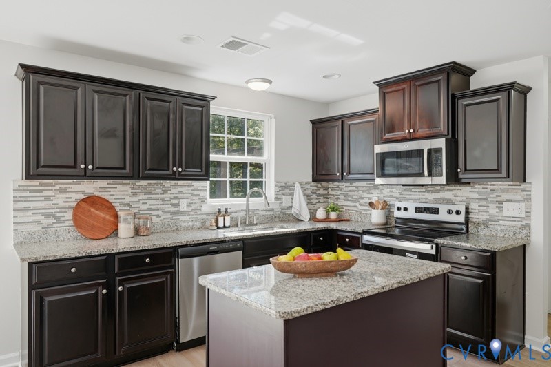Kitchen featuring dark wood-finish cabinetry, light speckled countertops, a tile backsplash, stainless steel appliances, and a central island