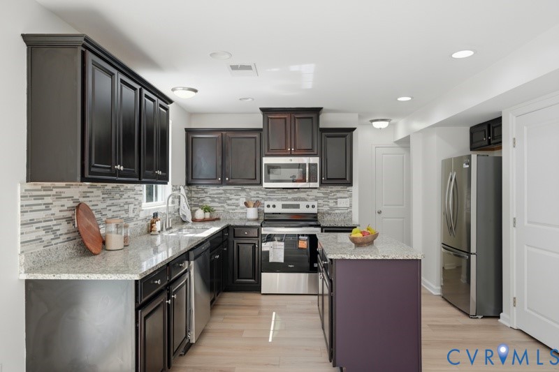 Kitchen featuring dark wood cabinetry, light-toned granite countertops, a mosaic tile backsplash, stainless steel appliances, and a central island with a contrasting base