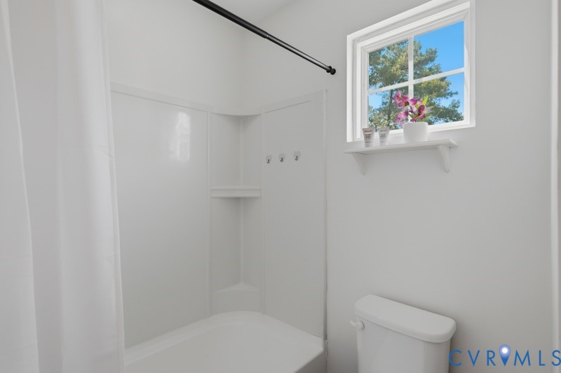 White tub and shower combination with an integrated soap dish, a white-framed window, and a wall-mounted toilet