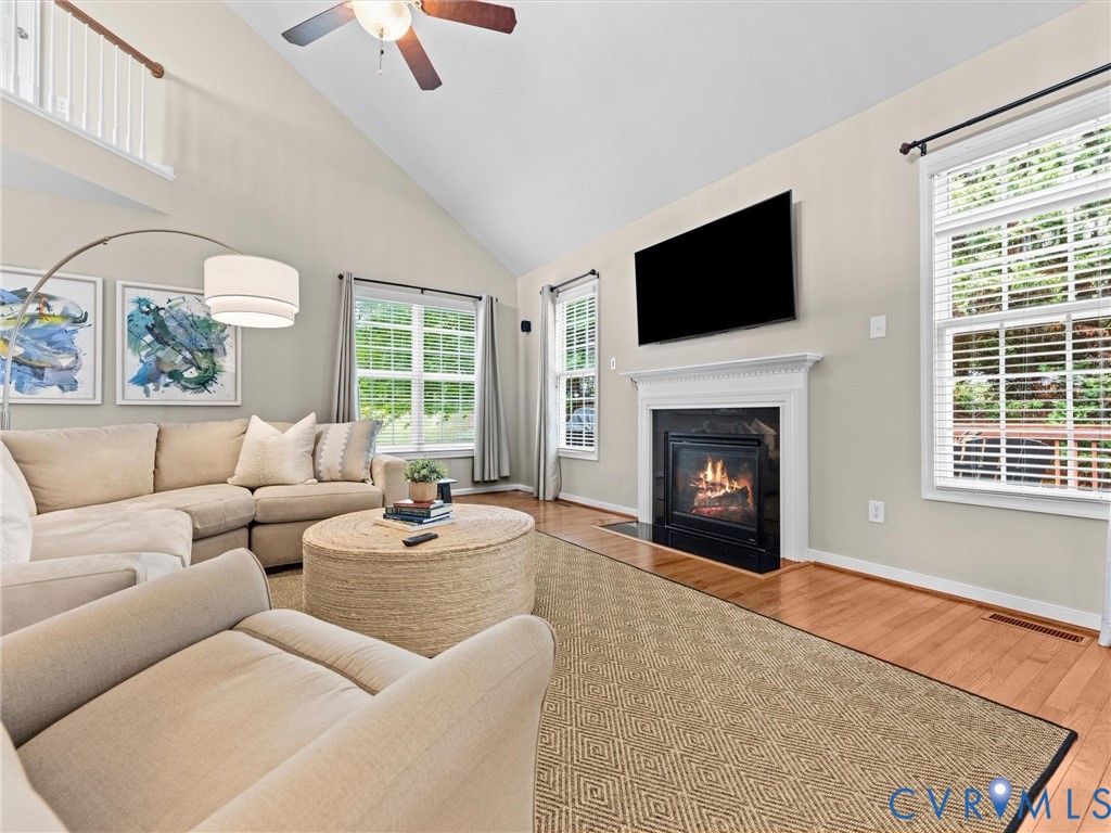 Vaulted ceiling, hardwood flooring, fireplace with white mantel, two large windows, and a ceiling fan