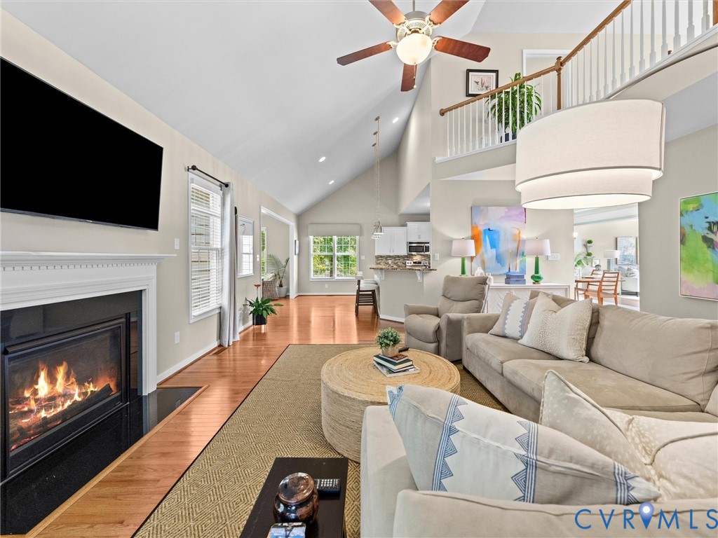 Vaulted ceiling with ceiling fan, wood-finish flooring, white mantel fireplace with black surround, white staircase with wood handrail, and an upper-level balcony