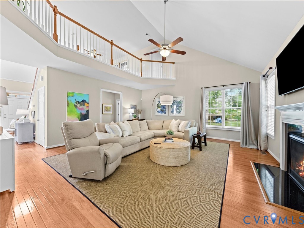 Vaulted ceiling with a ceiling fan, wood-finish flooring, two-story living area, fireplace with a white mantel, and an upper-level railing overlooking the main living space