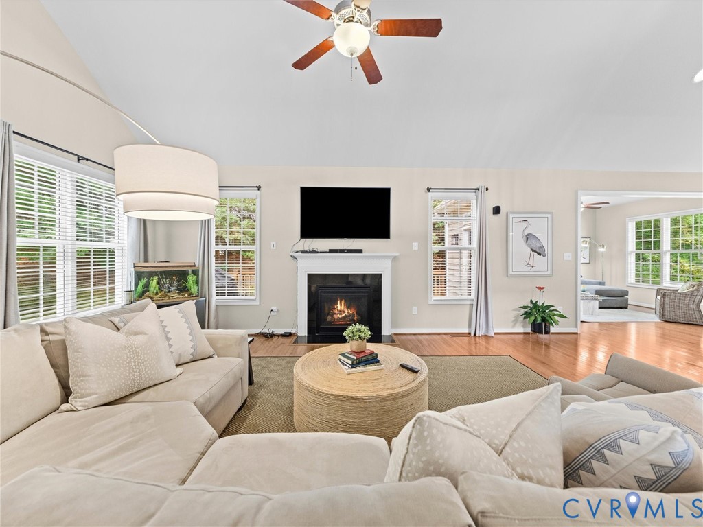 Vaulted ceiling with a ceiling fan, a fireplace with a dark surround, multiple windows with blinds, and wood-finish flooring