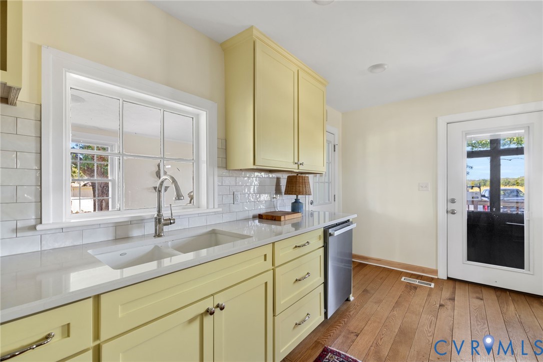 Kitchen featuring wood-finish flooring, light yellow cabinetry, undermount double-basin sink, stainless steel dishwasher, and white subway tile backsplash
