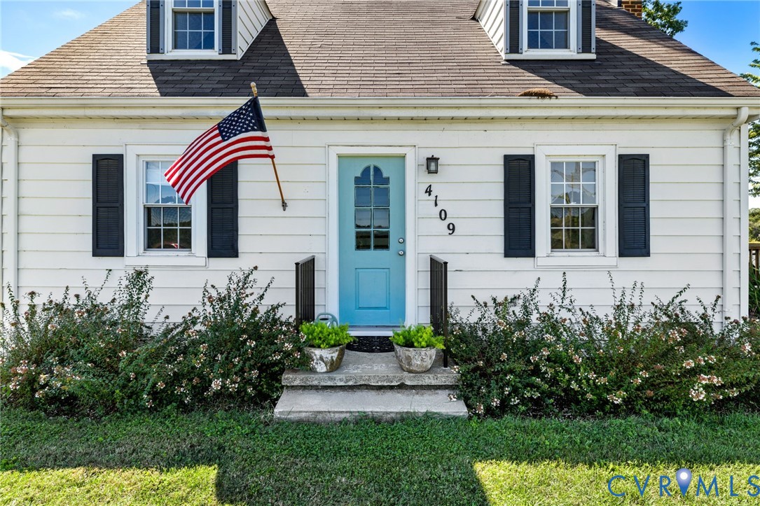 White clapboard siding with a light blue front door, dormer windows, dark window shutters, and a stone front step