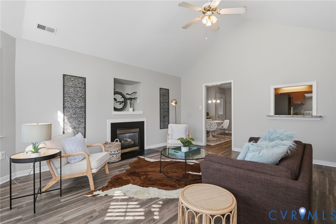 Vaulted ceiling with a ceiling fan, fireplace with a mantel and built-in niche, wood-finish flooring, and a pass-through window with a countertop Vaulted ceiling with a ceiling fan, fireplace with a mantel and built-in niche, wood-finish flooring, and a pass-through window with a countertop