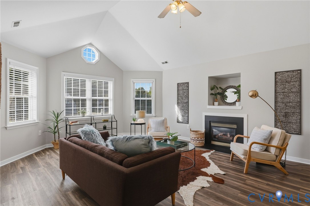 Vaulted ceiling with ceiling fan, prominent octagonal window, white window blinds, wood-finish flooring, and a built-in fireplace with mantel Vaulted ceiling with ceiling fan, prominent octagonal window, white window blinds, wood-finish flooring, and a built-in fireplace with mantel