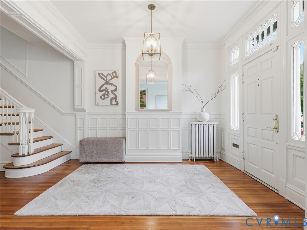 Hardwood flooring, paneled walls, ornate front entry door with transom and sidelights, staircase with wood treads, and a geometric chandelier Hardwood flooring, paneled walls, ornate front entry door with transom and sidelights, staircase with wood treads, and a geometric chandelier