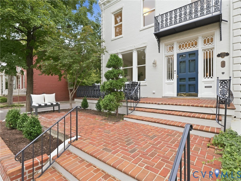 White facade with a blue paneled entry door, ornate iron balcony, brick paver courtyard, and wrought iron railings White facade with a blue paneled entry door, ornate iron balcony, brick paver courtyard, and wrought iron railings