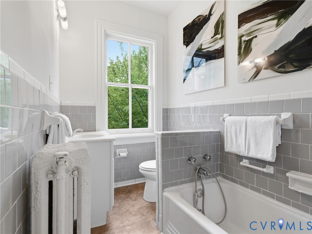 Bathroom with white subway tile and gray square tile wainscoting, featuring a built-in bathtub with chrome fixtures, a window, and a white pedestal sink