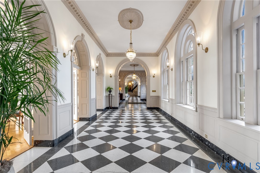 Grand gallery hall featuring arched entryways, black-and-white checkered stone flooring, decorative crown molding with ceiling medallions, dual-tone wainscoting, and crystal chandeliers