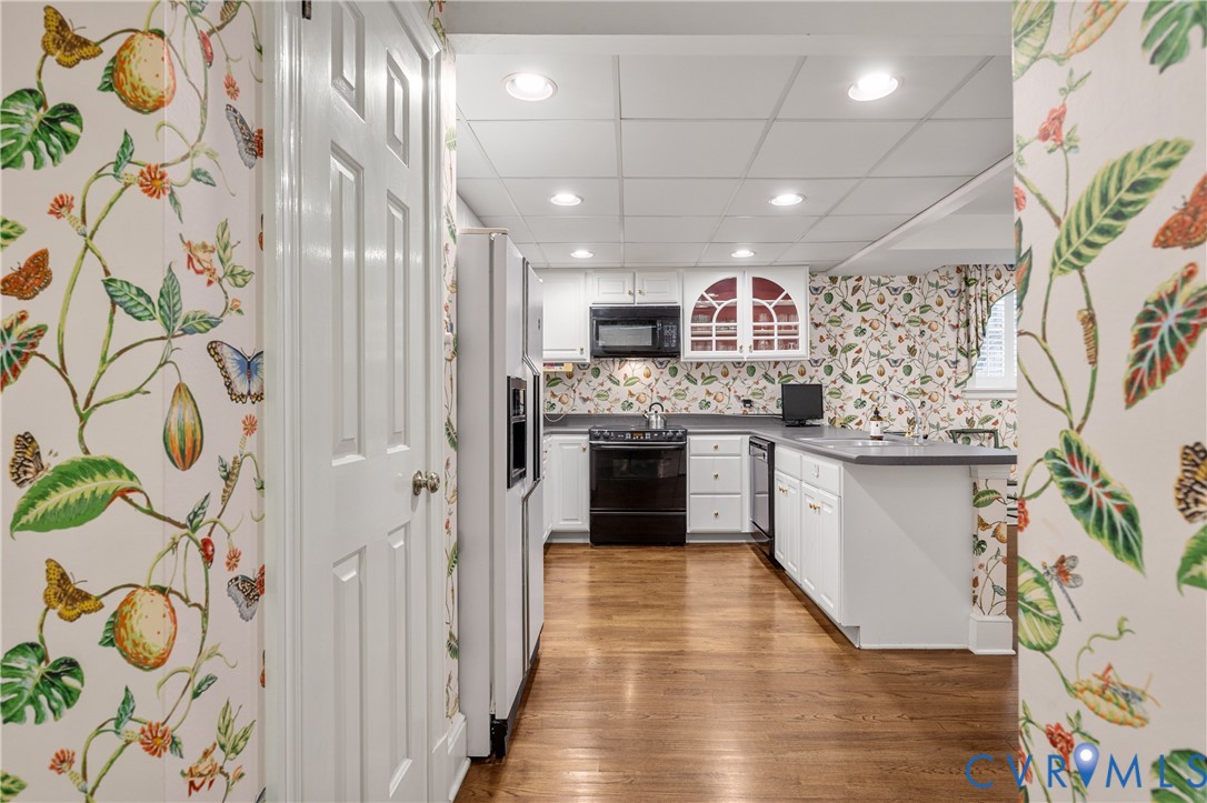 Kitchen featuring wood-finish flooring, white cabinetry with glass-front display accents, integrated black appliances, and a peninsula with countertop space