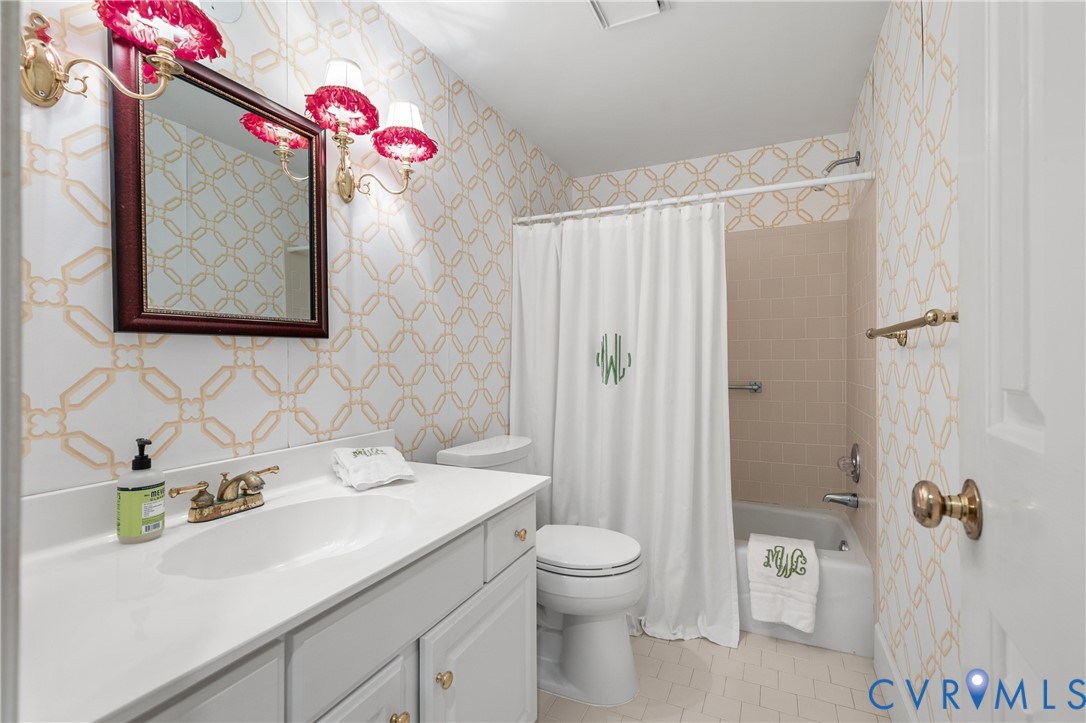 Bathroom featuring a vanity with a white solid surface countertop, brass-finish faucet hardware, patterned wallcovering, dual brass-finish wall sconces, and a tiled shower surround