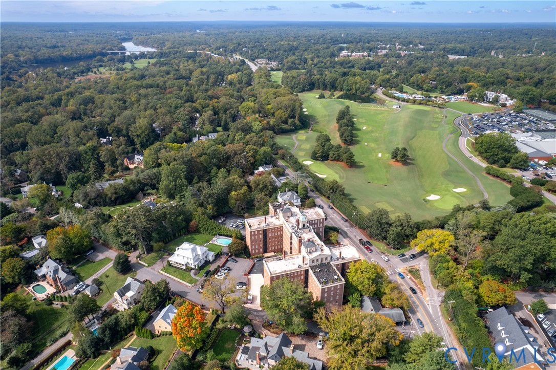 Aerial perspective of a multi-story brick building situated amidst extensive wooded acreage, featuring proximity to an expansive golf course and surrounding mature tree canopy