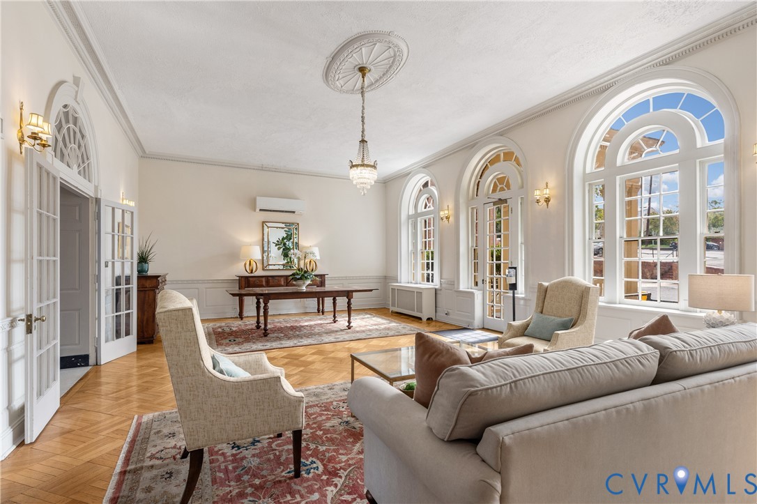 Living space featuring herringbone-patterned wood flooring, arched windows with fanlight detailing, crown molding, wall paneling, and an ornate ceiling medallion with a crystal chandelier