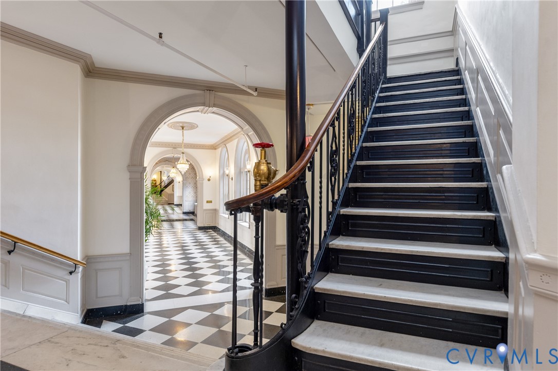 Staircase featuring black iron railings, a wood handrail, and stone-tread steps, complemented by arched entryways, crown molding, and a checkered stone floor pattern