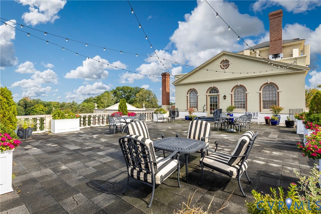 Expansive rooftop terrace featuring stone paver flooring, a classic stone balustrade, two brick chimneys, and overhead string lighting