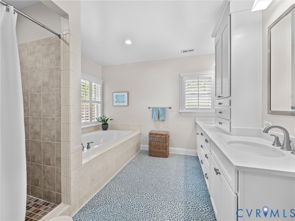 Tile-surround soaking tub, dual vanity with white cabinetry, tile shower with mosaic floor, window with plantation shutters, and recessed lighting