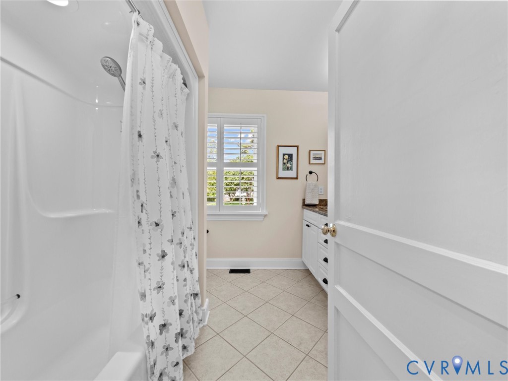 Ceramic tile flooring, white shaker-style vanity with dark hardware, granite countertop, and a window with plantation shutters