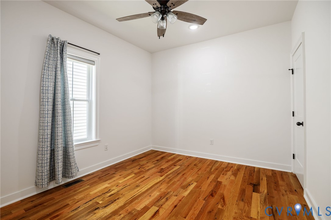 Room featuring wood-finish flooring, a multi-blade ceiling fan with integrated lighting, and a single-hung window with a roller shade