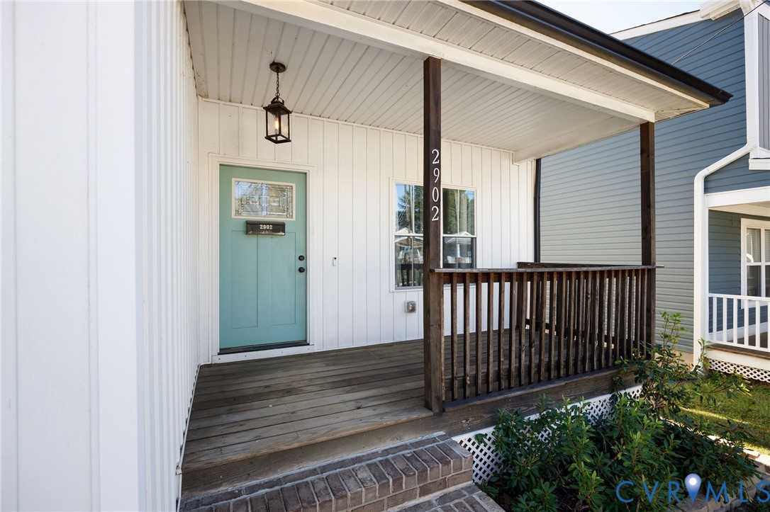 Covered front porch with wood decking, vertical board-and-batten siding, dark wood railing, brick-stepped entrance, and a decorative paneled door