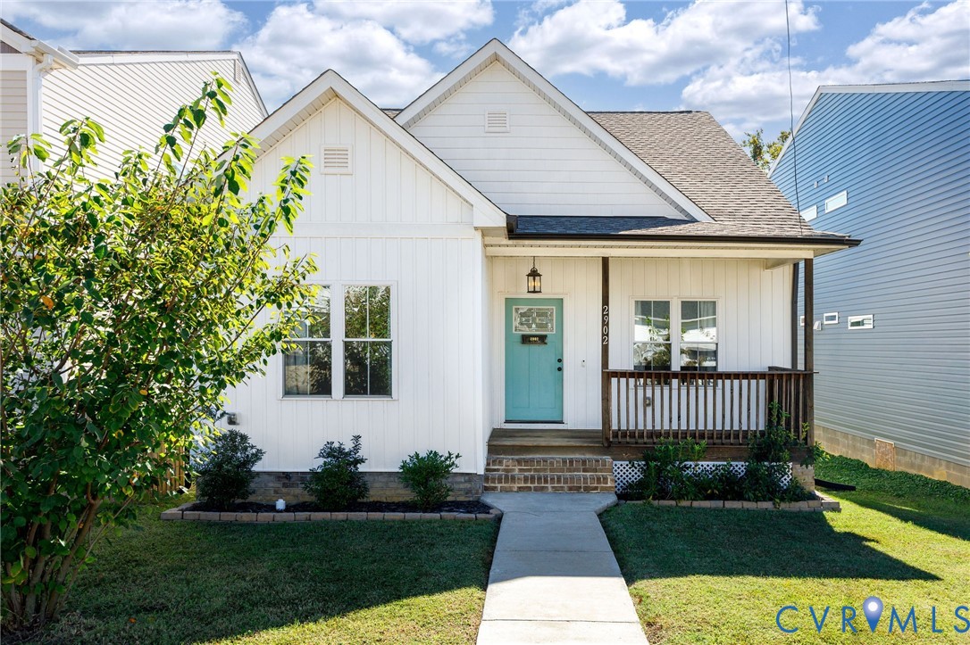Single-story residence featuring white vertical siding, a covered front porch with a dark wood-finish railing, a brick-accented entryway, and a solid-color front door Single-story residence featuring white vertical siding, a covered front porch with a dark wood-finish railing, a brick-accented entryway, and a solid-color front door