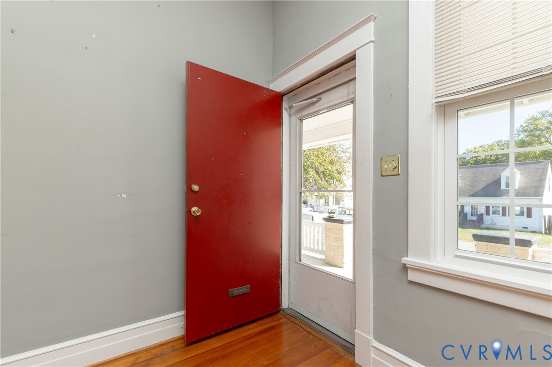 Entryway featuring wood-finish flooring, a solid red door with integrated mail slot, and a multi-pane window with louvered blinds
