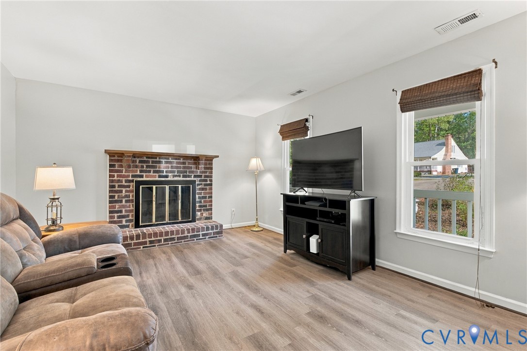 Brick fireplace with a wood mantel, wood-finish flooring, and multiple windows with integrated window treatments