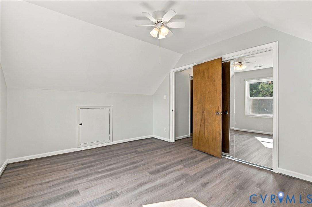 Sloped ceiling design with wood-finish flooring, mirrored closet doors, built-in storage access panel, and a multi-blade ceiling fan with integrated lighting