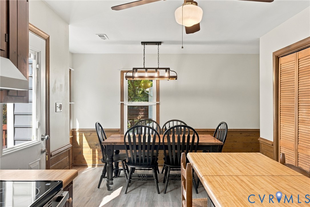 Dining area featuring wood-plank wainscoting, linear multi-bulb pendant lighting, wood-finish flooring, a ceiling fan, and bi-fold louvered closet doors