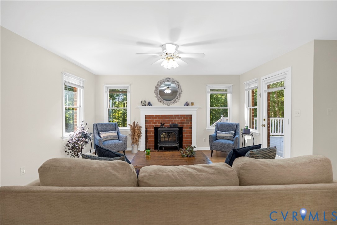 Living area featuring a brick fireplace with a wood-burning insert and white mantel, large windows, wood-finish flooring, and a ceiling fan with integrated lighting