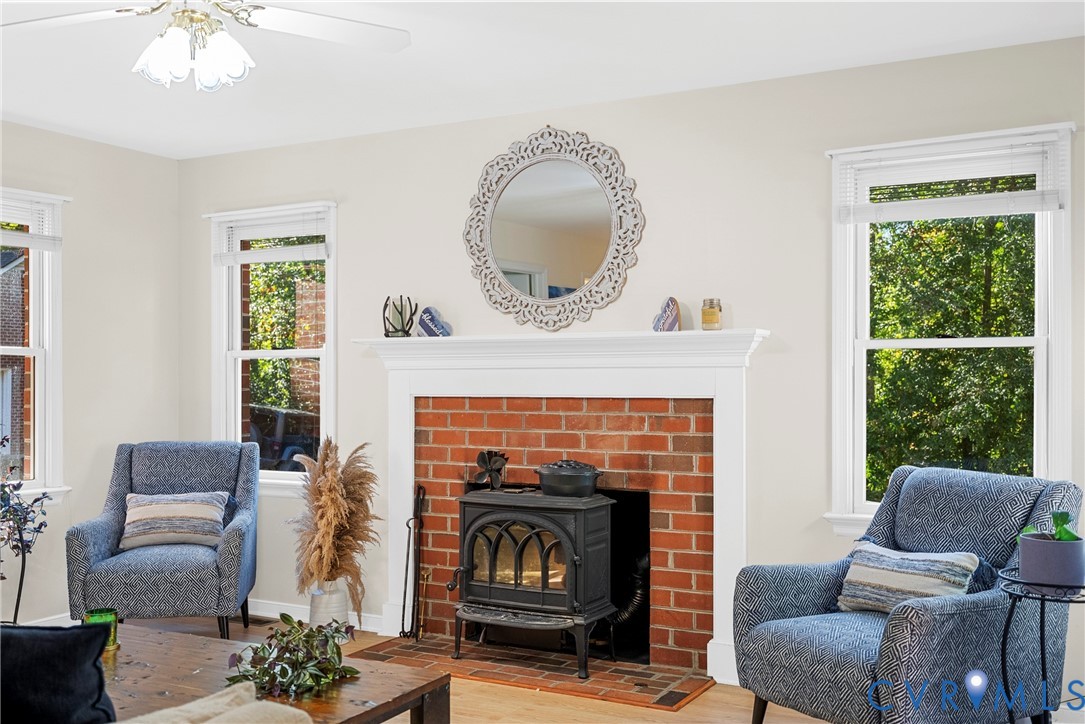 Exposed brick fireplace featuring a cast-iron wood-burning stove, white mantel, and wood-finish flooring, illuminated by natural light from multiple double-hung windows