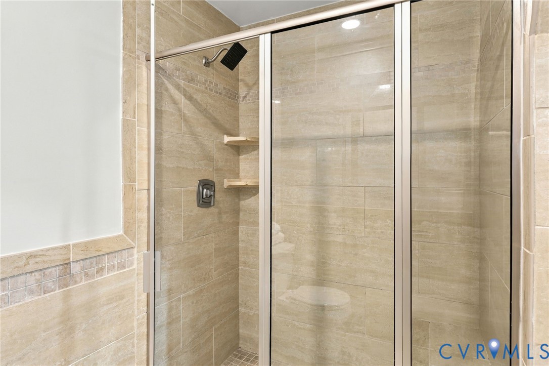 Walk-in shower featuring floor-to-ceiling neutral tile, semi-frameless glass enclosure, matte black rainfall showerhead, integrated corner shelving, and mosaic tile accents