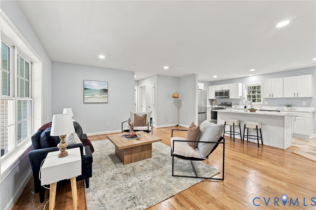 Open-concept area featuring wood-finish flooring, recessed lighting, and a kitchen equipped with white cabinetry, a center island with bar seating, and stainless steel appliances Open-concept area featuring wood-finish flooring, recessed lighting, and a kitchen equipped with white cabinetry, a center island with bar seating, and stainless steel appliances
