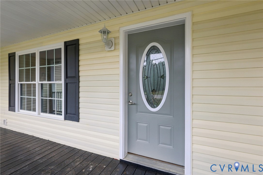 Covered front porch with dark-toned decking, light-colored horizontal siding, dark window shutters, and a front entry door featuring an oval decorative glass insert Covered front porch with dark-toned decking, light-colored horizontal siding, dark window shutters, and a front entry door featuring an oval decorative glass insert