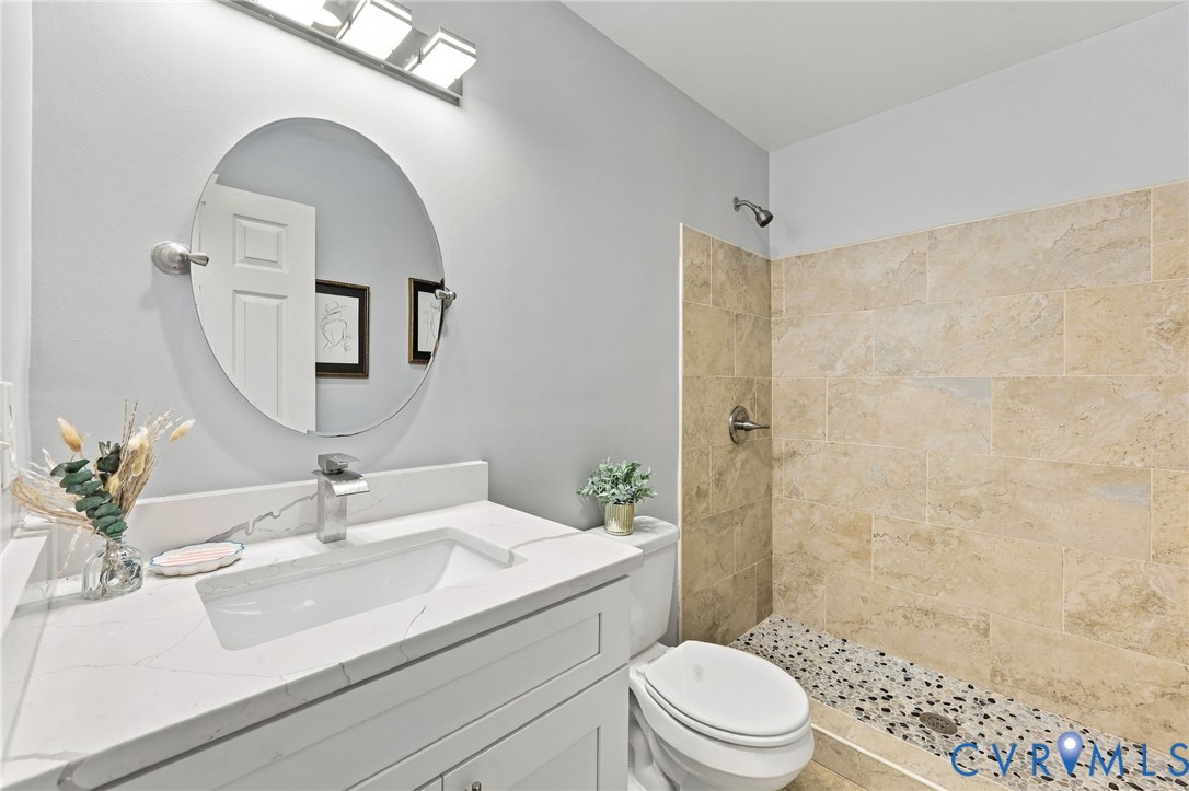 Bathroom vanity with white stone-finish countertop, undermount sink, and oval mirror, accompanied by a walk-in shower featuring textured stone tile walls and a pebble-stone floor