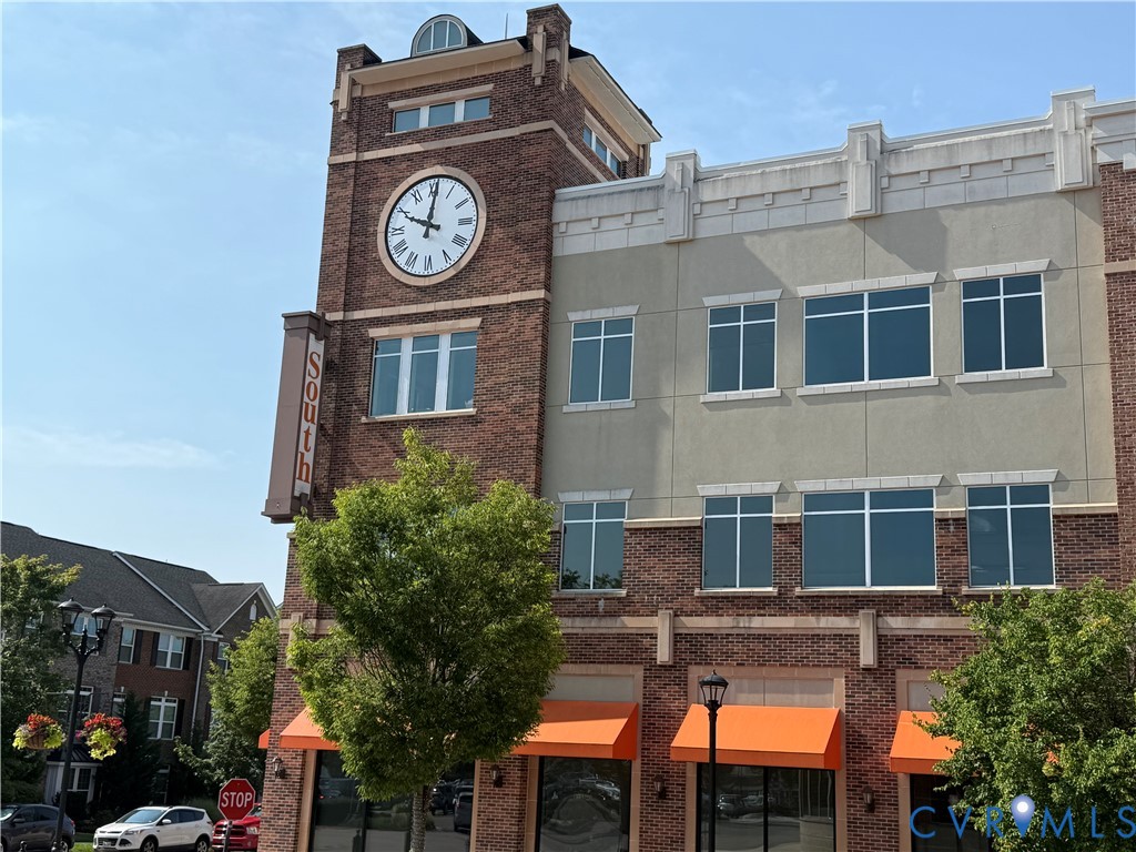 Historic brick mixed-use building featuring a prominent clock tower with illuminated dial, large storefront windows with orange awnings on the ground level, and a contemporary tan faade addition