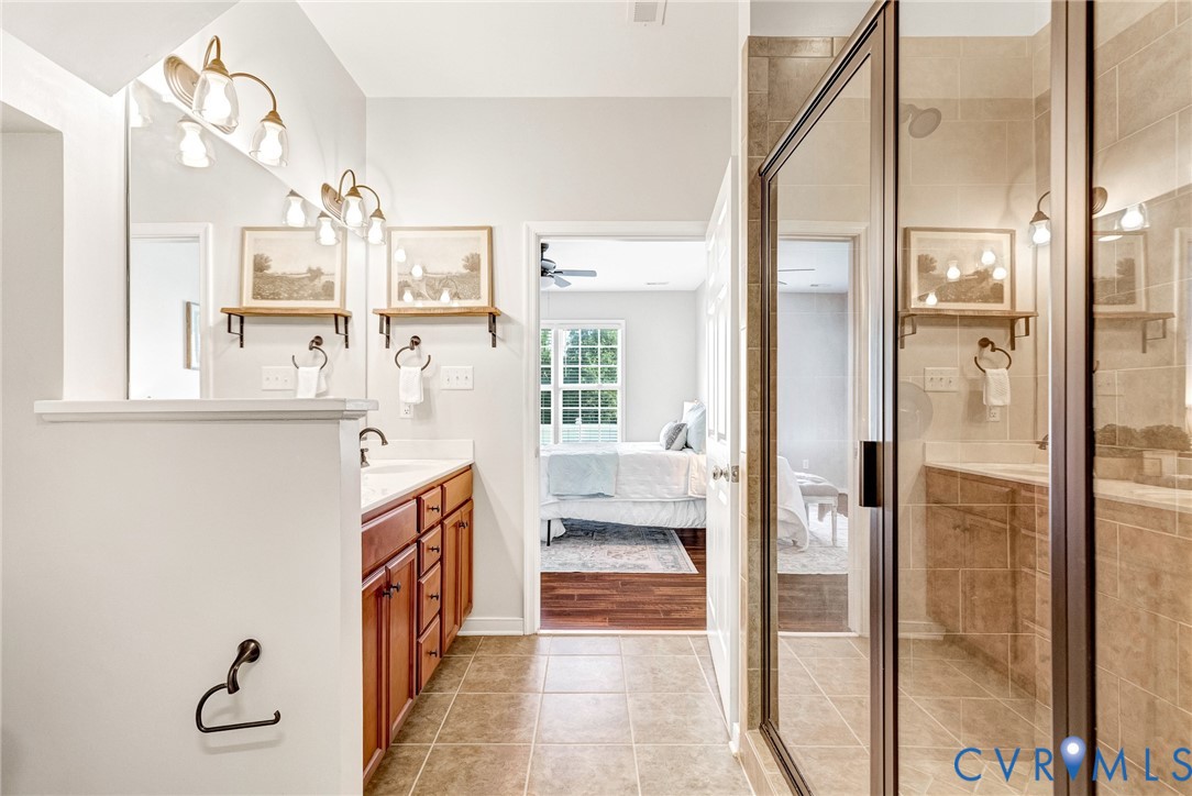 Dual vanity with wood cabinetry and undermount sinks, bronze-finish wall sconces, large wall-mounted mirror, and a tiled walk-in shower with a glass enclosure