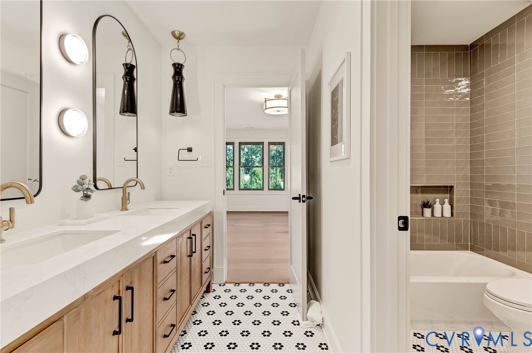Dual-vanity cabinet with wood-finish cabinetry, quartz countertops, arched mirrors with integrated lighting, matte gold-finish hardware, and geometric patterned floor tiling