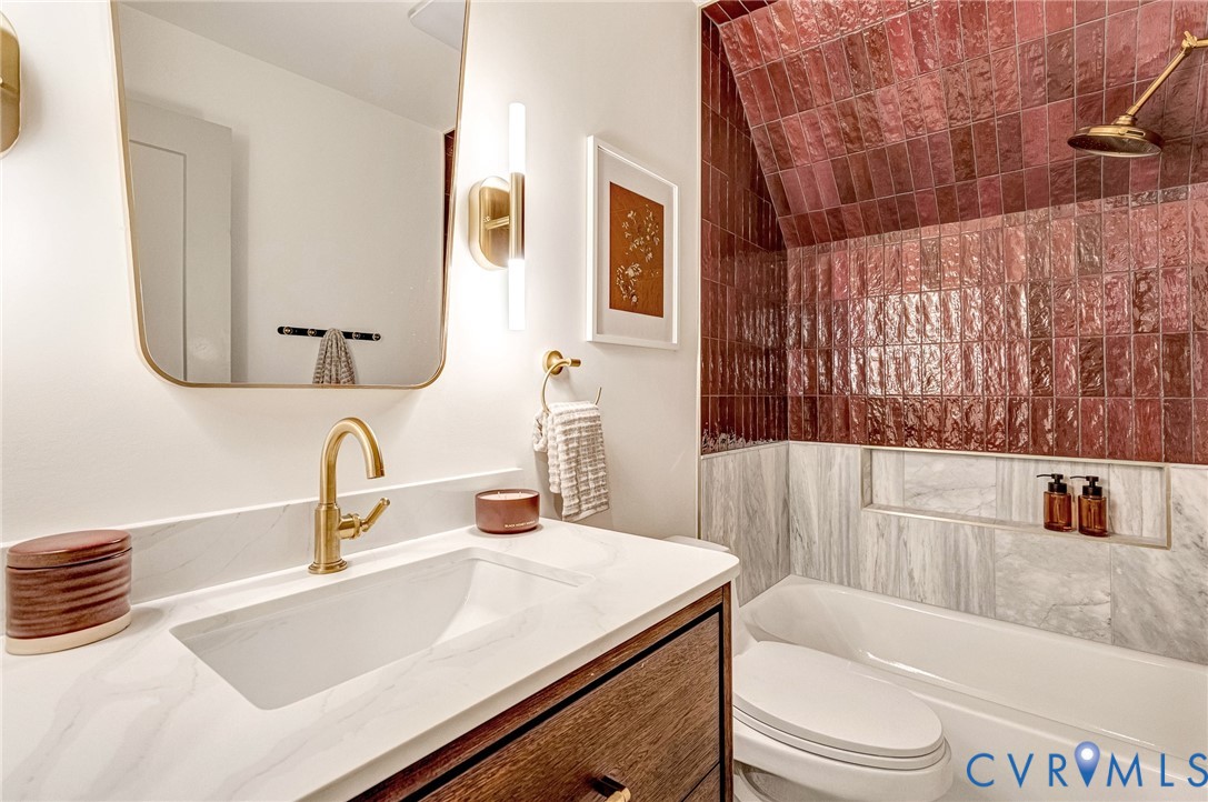 Bathroom featuring a wood-finish vanity with a quartz countertop, brass-finish hardware, a wall-mounted rectangular mirror, and a shower area with glazed burgundy tile and stone-finish wall tile
